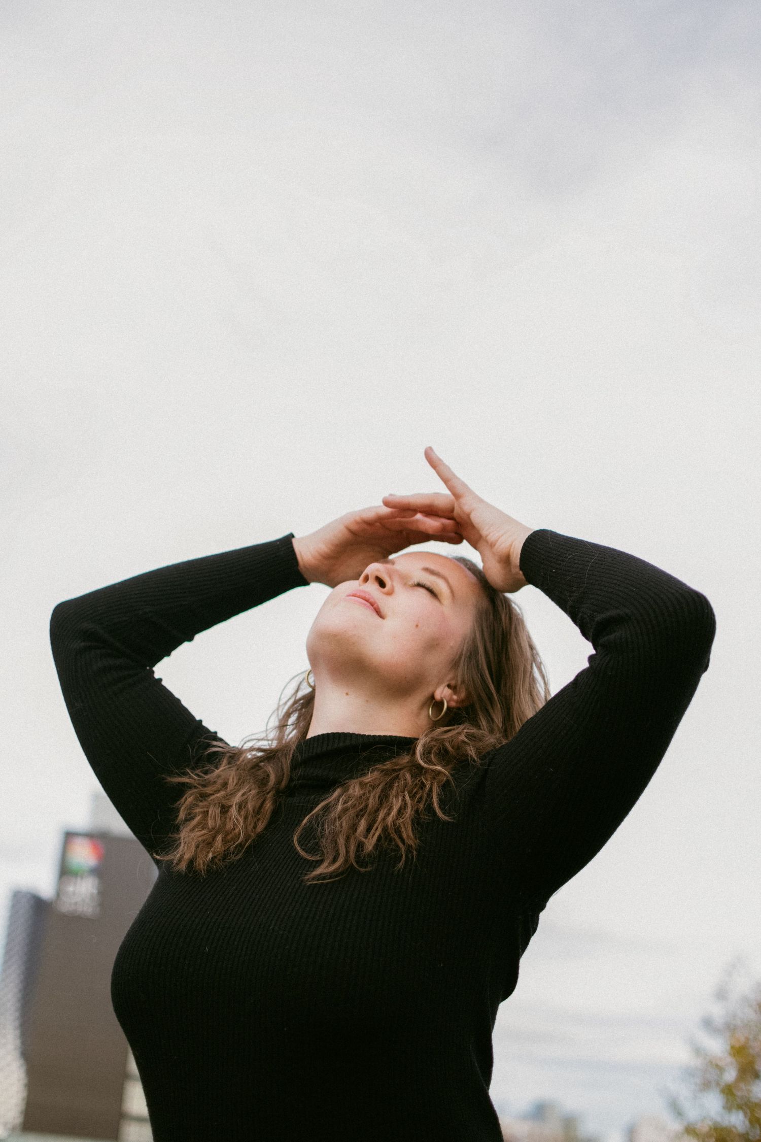 A dramatic portrait of someone in black sweater posing with arms raised against a cloudy sky.