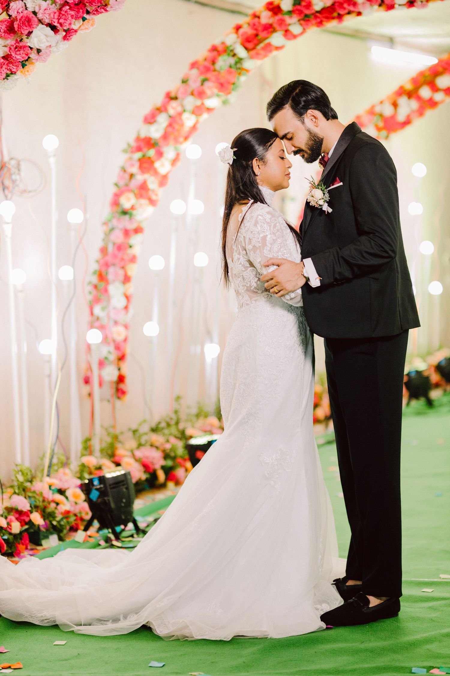Wedding couple shares romantic moments under an arch decorated with pink and red flowers on a green carpet.