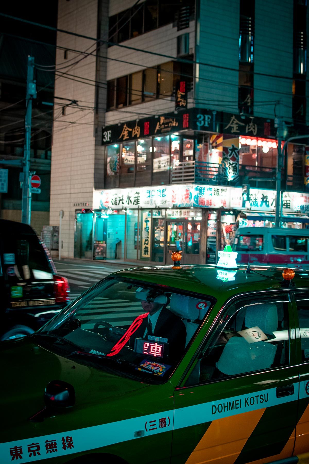 Green taxi cab parked outside brightly lit Japanese storefront at night.
