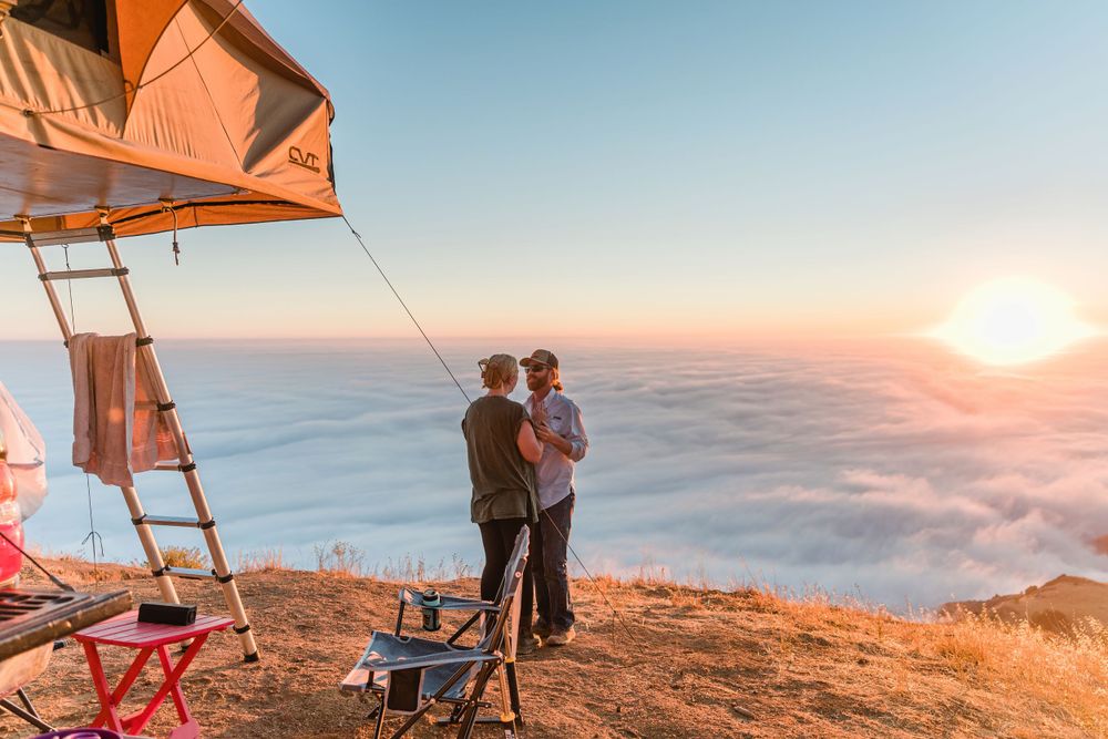 Couple watching sunset from mountain overlook with rooftop tent camping setup.