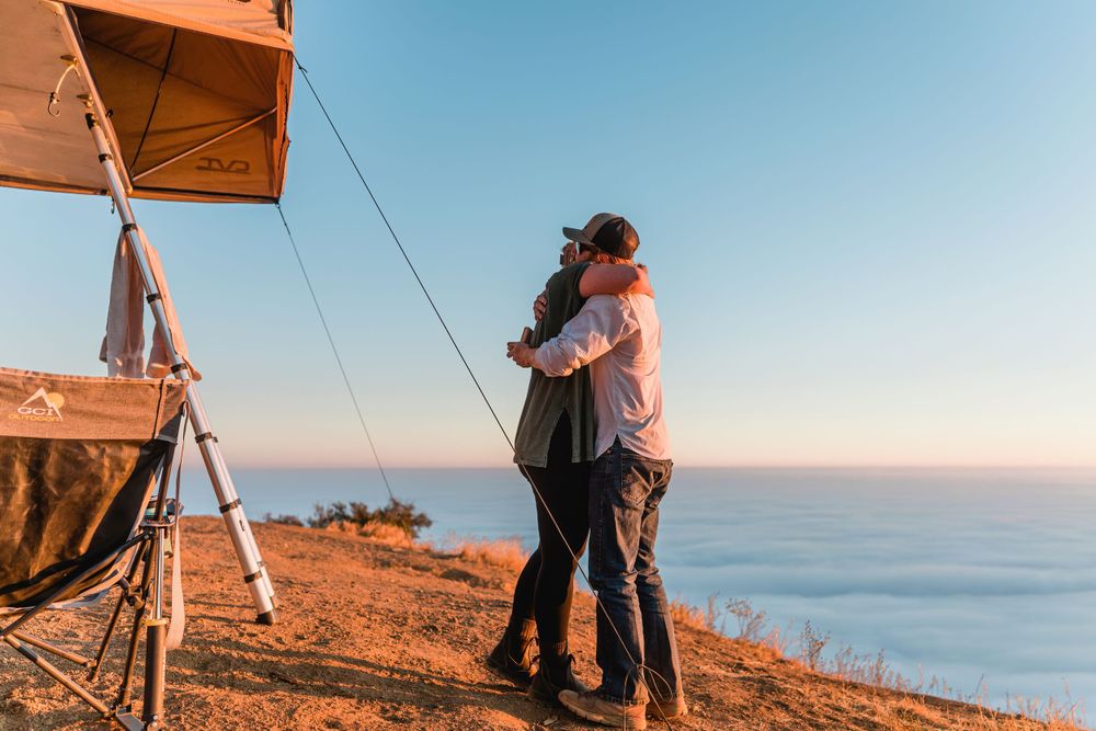 Couple embracing on clifftop during sunset with rooftop tent camping setup overlooking ocean views.