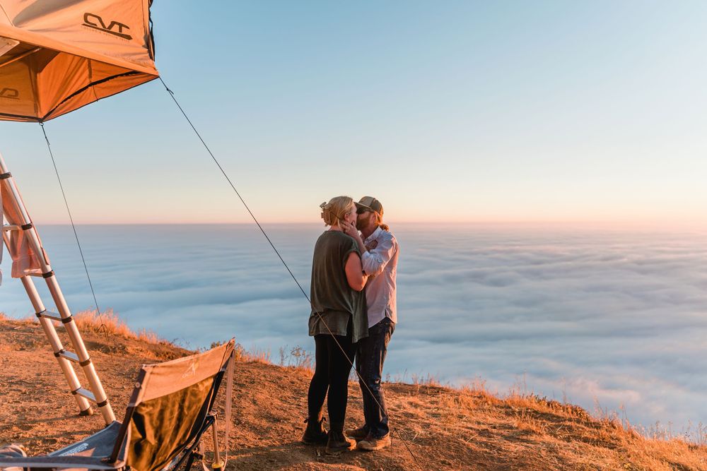 Silhouetted couple embracing on cliff edge during golden hour with rooftop tent setup.