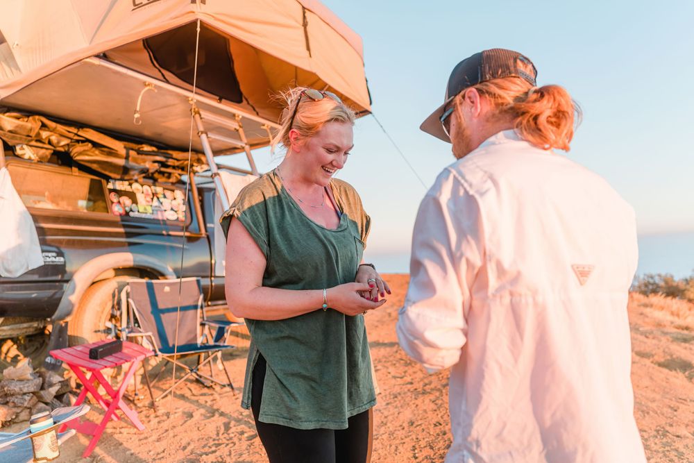 Camping scene with rooftop tent and people gathered on scenic cliff during golden hour.