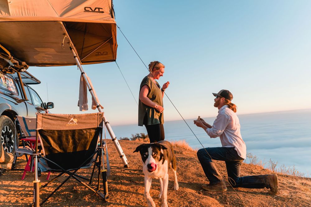 Campers and their dog enjoying scenic overlook from their rooftop tent setup above clouds at sunset.