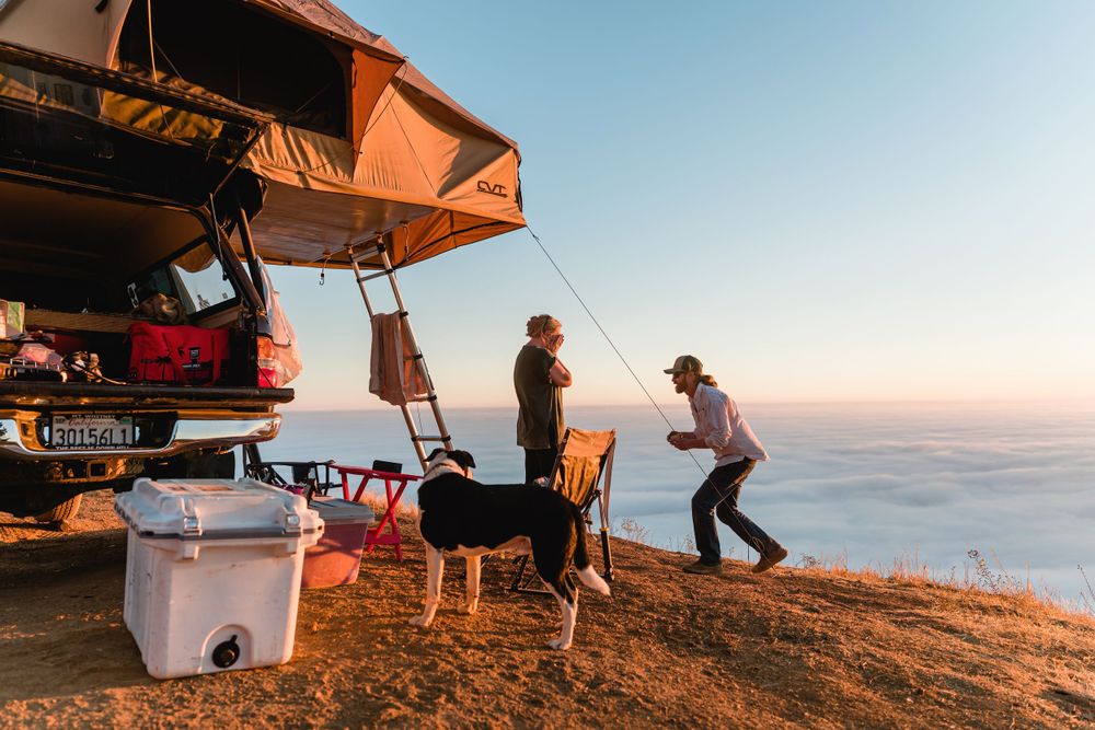 People camping on a cliff overlooking foggy ocean view with rooftop tent and cooking setup at sunset.