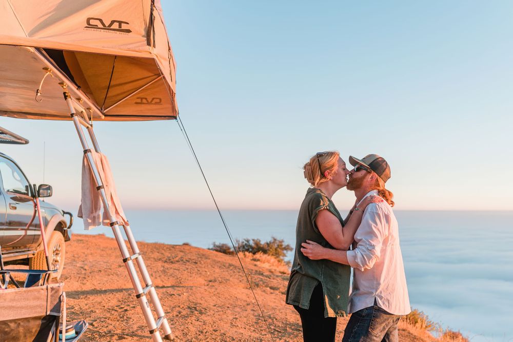Couple sharing intimate moment at sunset while camping with rooftop tent on coastal cliff.
