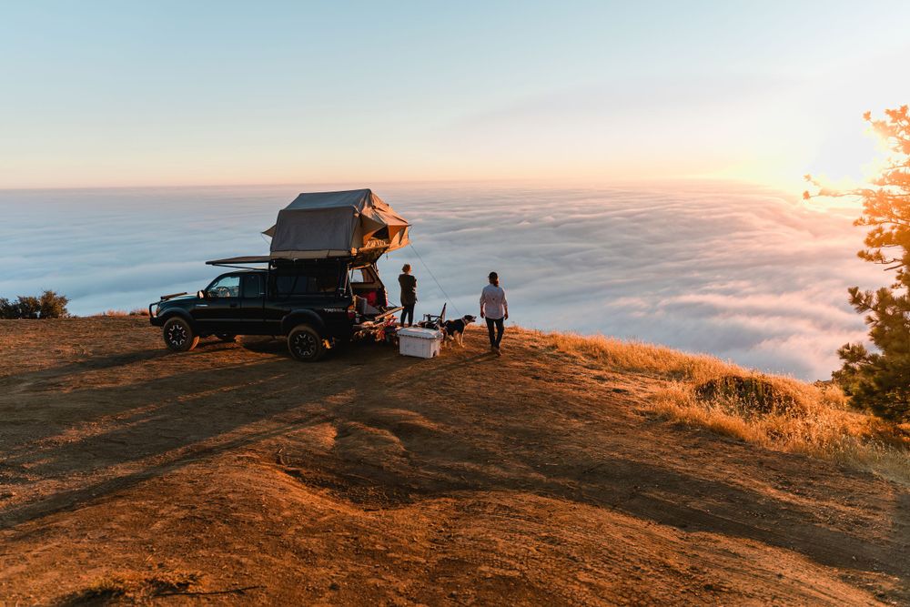 Car with rooftop tent parked on cliff edge overlooking foggy ocean at sunset.