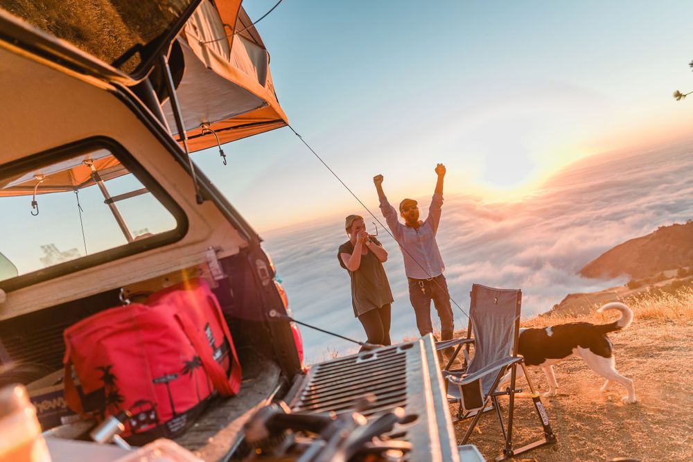Campers celebrating beautiful sunset view from their rooftop tent setup on mountain overlook.