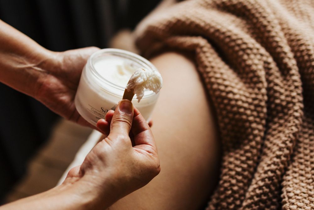 Hands scooping white cream from jar during spa treatment.