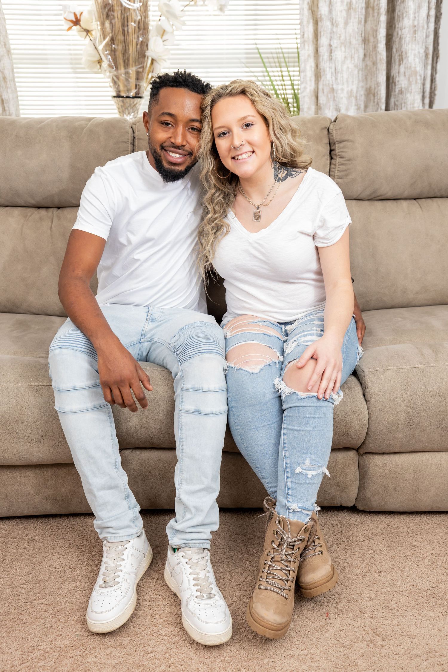 Couple in casual ripped jeans and white t-shirts sit close together on beige couch.