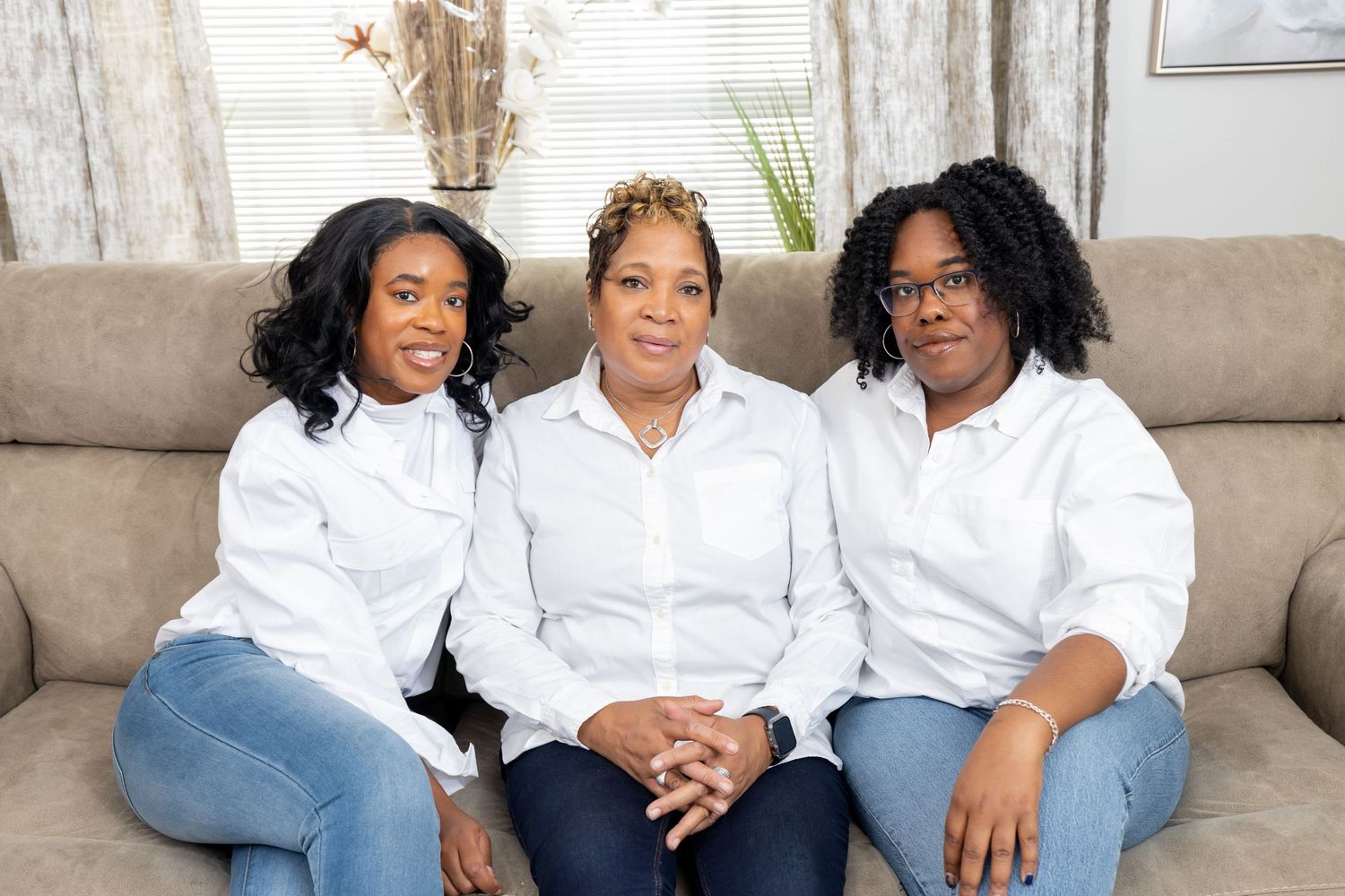 Three people wearing coordinated white shirts and jeans smile while seated on beige couch.
