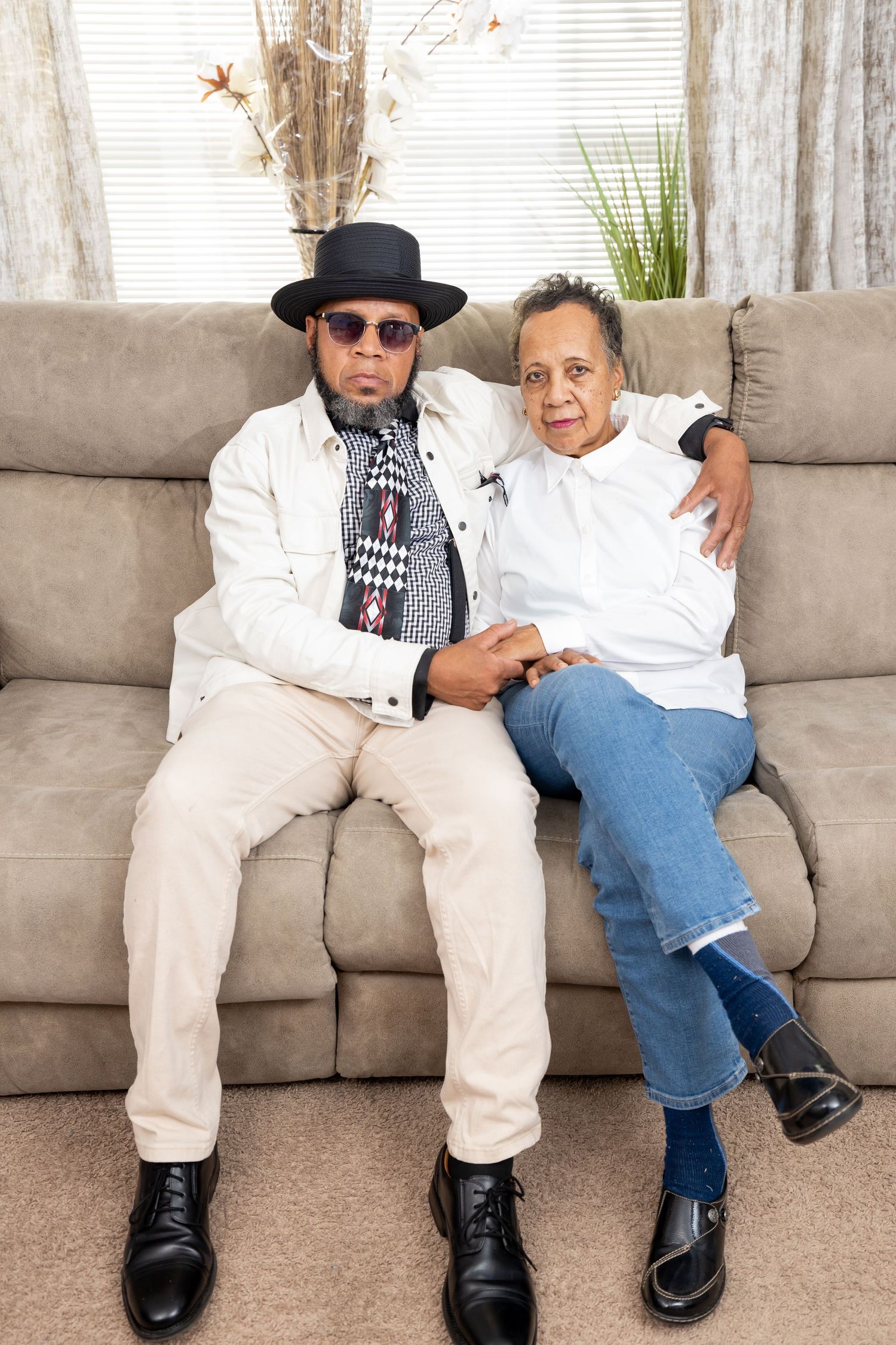 A couple relaxes on a beige couch, with one wearing sunglasses and a hat while dressed in light colored clothing.