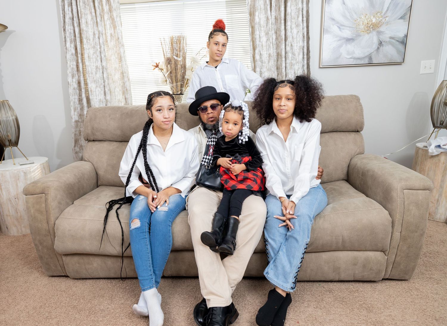 Group photo on beige couch with someone in sunglasses and hat centered among others in white shirts and jeans.