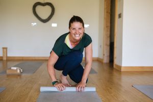 Person in dark shirt rolling out a yoga mat in a bright exercise room with wooden floors.