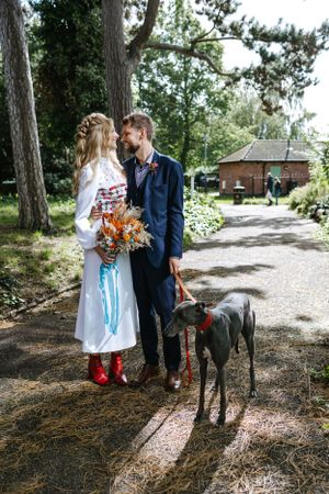 A couple poses on a garden path with their greyhound dog and orange floral bouquet.