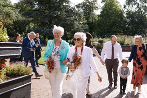 Wedding guests walking through a sunny garden path carrying flowers.