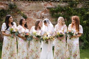Bridesmaids in floral print dresses holding bouquets pose against rustic brick wall.