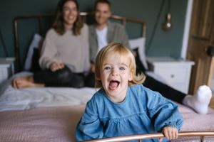 Excited child in blue dress sits on bed while parents watch from behind.