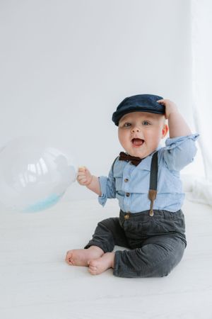 Stylishly dressed infant playing with a bubble while sitting on white floor.