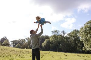 Adult lifting child high into air against backdrop of sunny meadow and trees.