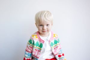 Small child wearing colorful holiday sweater with festive patterns sits against white background.