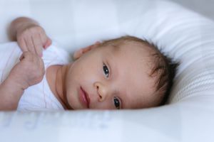 A baby lying on white bedding gazes at the camera with bright eyes.