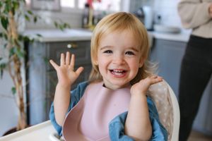 Happy toddler in pink bib waves and smiles brightly in kitchen setting.