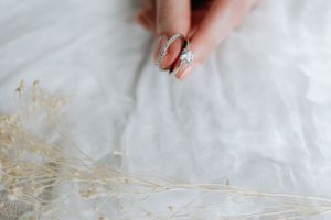 Close-up of hand with engagement ring next to dried flowers on white fabric.