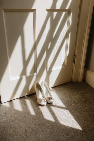 White bridal heels sit in dramatic window light casting geometric shadows on a carpeted floor.