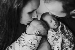 Black and white intimate portrait of parents with sleeping newborn twins in floral outfits.