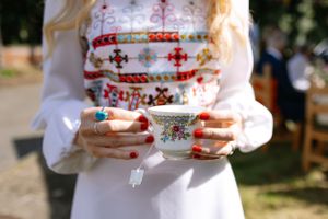 Hands hold a delicate teacup with floral embroidered dress detail visible.