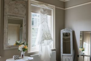 A wedding dress hangs elegantly in front of a bright window with vintage mirrors and floral decor.