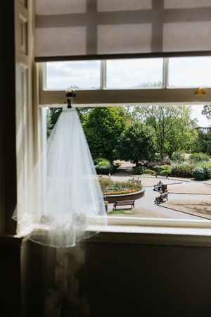 A delicate white wedding veil hangs in a window overlooking a peaceful garden scene.