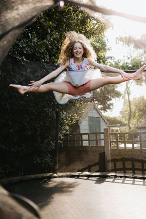 A dancer performs a dramatic split jump on a backyard trampoline with sunlight streaming through trees.