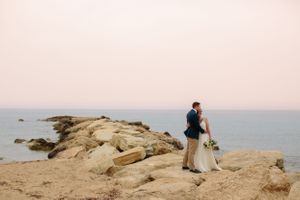 A couple stands together on rocky coastal cliffs overlooking the ocean at sunset.