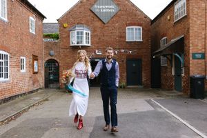 A couple walks together outside a brick building with red shoes and formal attire.
