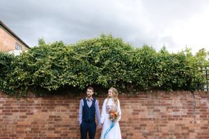A couple poses against a brick wall with green foliage overhead.