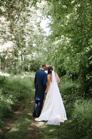A romantic wedding couple walking down a tree-lined garden path with the bride in white and groom in navy.