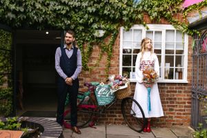 A couple poses by a vintage bicycle against an ivy-covered brick wall.