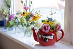 A red Yorkshire Tea teapot filled with colorful wildflowers sits on a sunny windowsill.