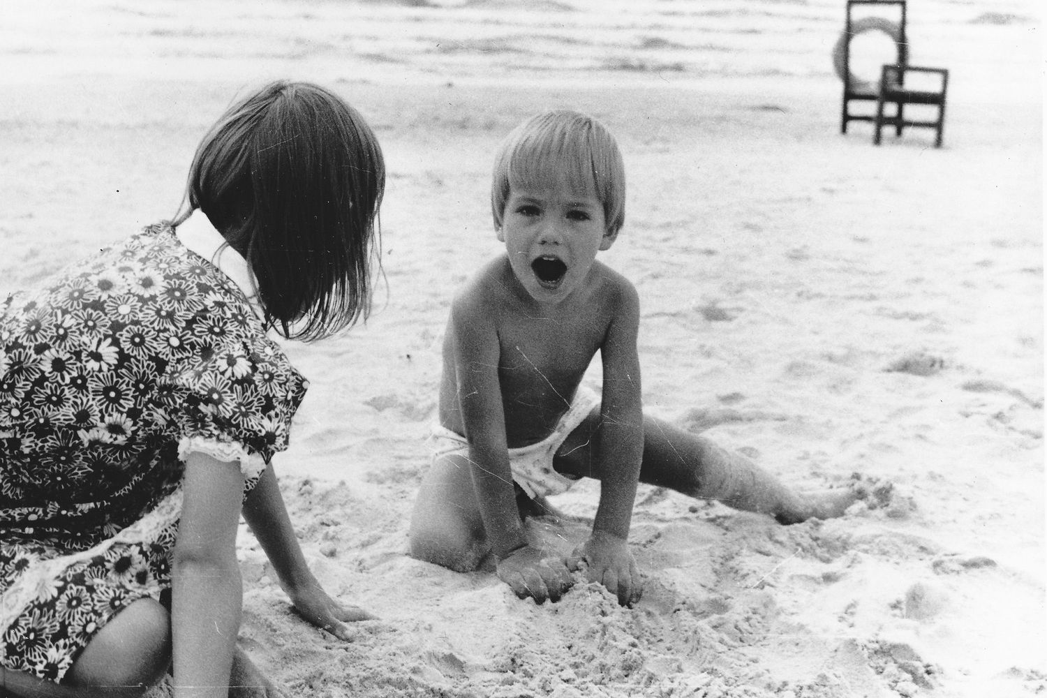 Black and white photograph of two kids playing in the sand on a beach with a chair in the background.