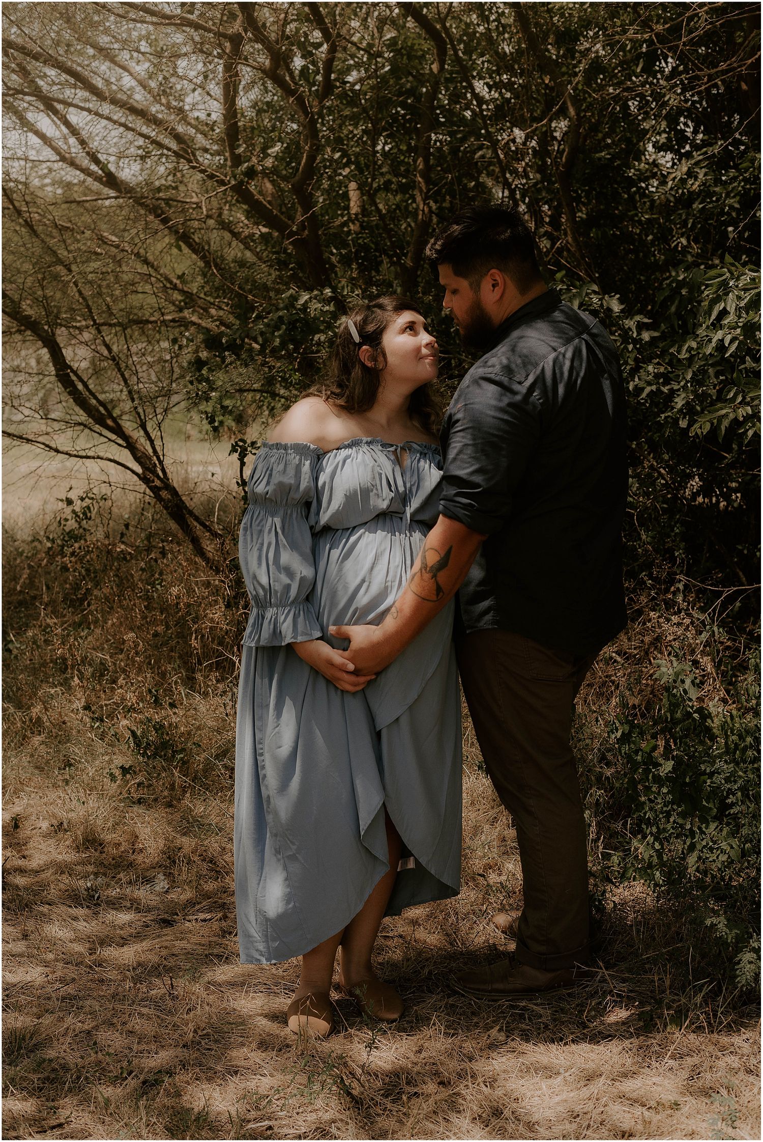Elegant maternity portrait featuring couple in casual attire against rustic outdoor backdrop.