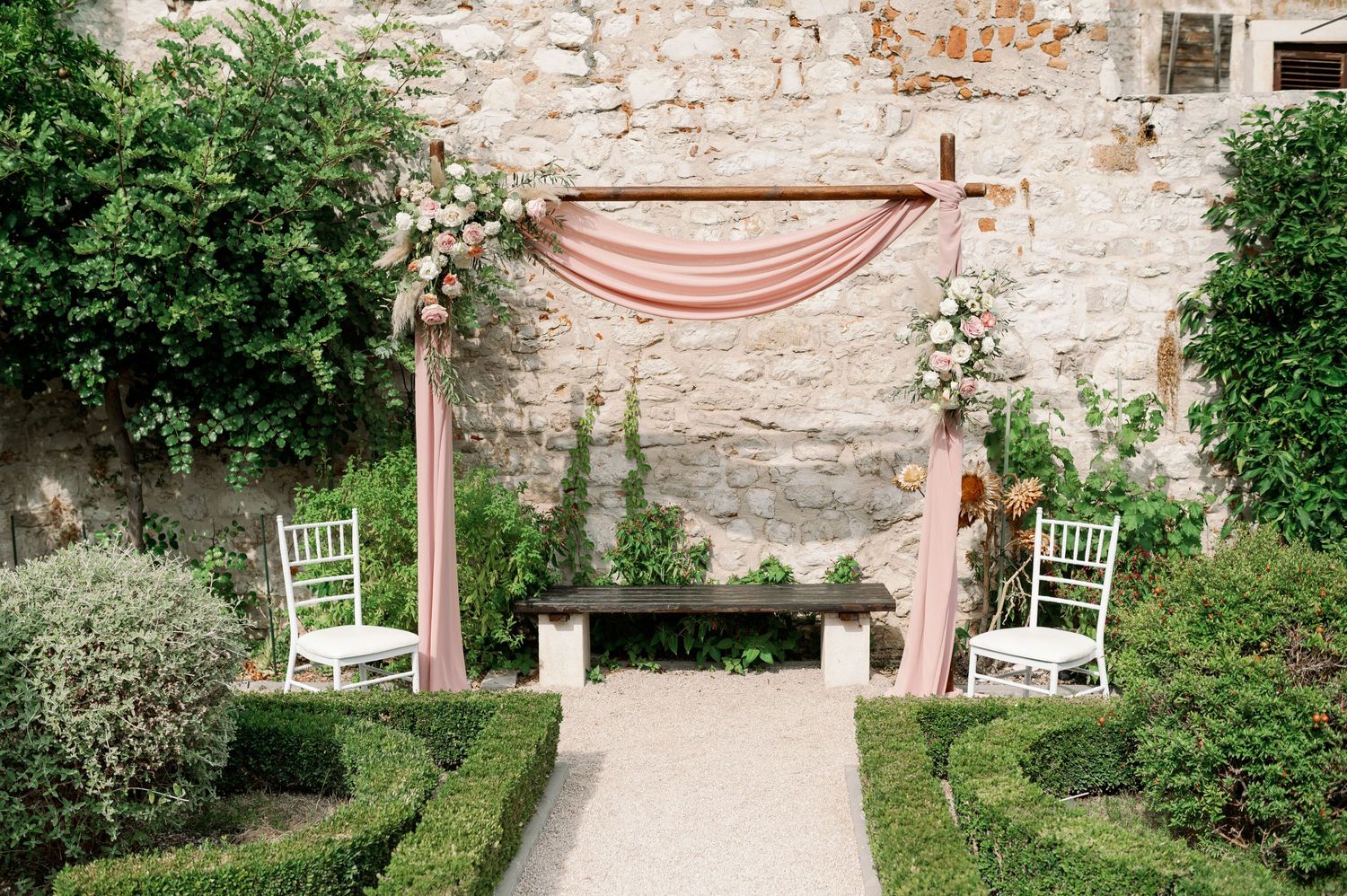 Romantic wedding ceremony setup with pink draping and white chairs against stone wall.