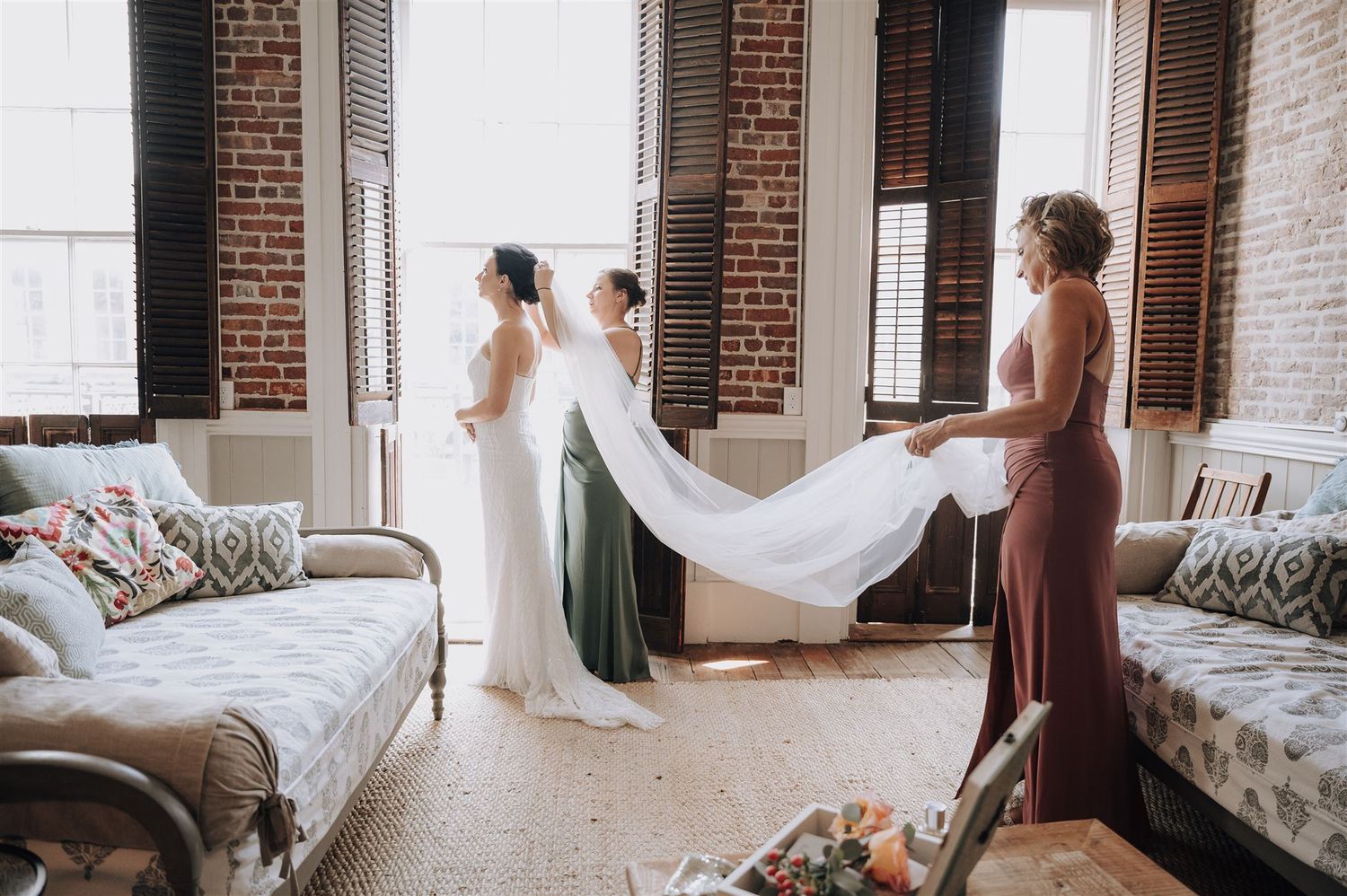 Bridesmaids in dusty rose dresses helping with the bride's veil in a sunlit room.