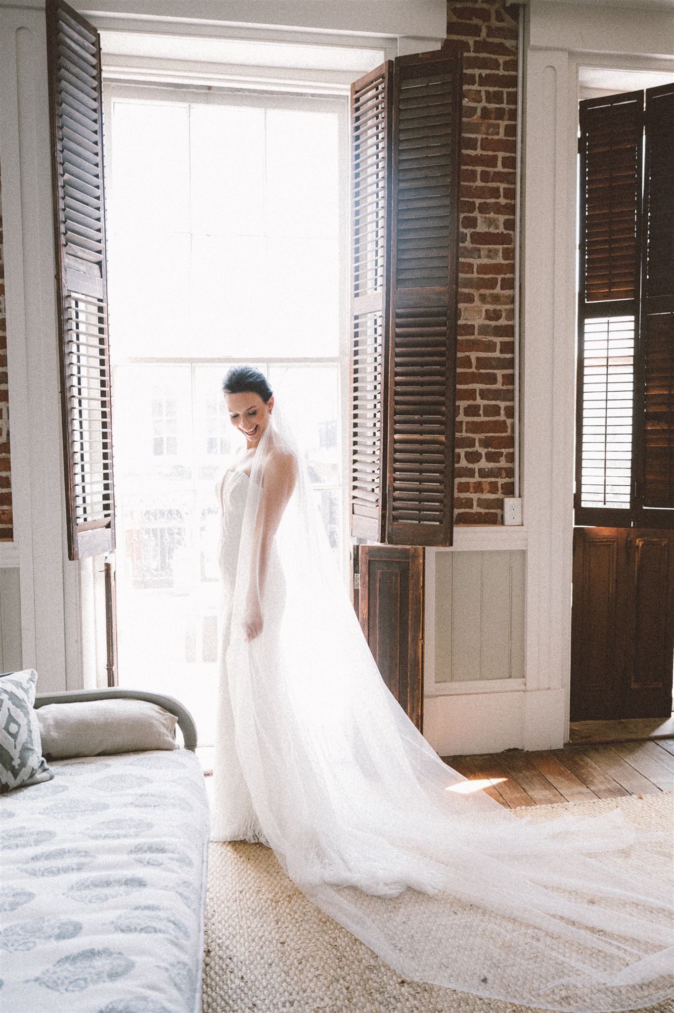 A bride in flowing white gown stands by vintage shuttered windows with natural light streaming in.