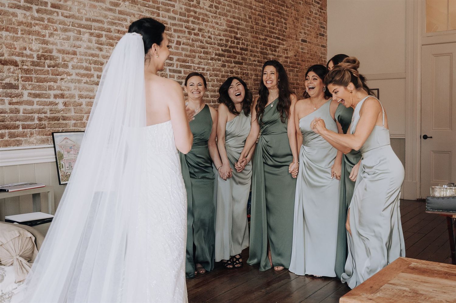 Wedding party in sage and silver dresses shares laughter with bride in rustic brick venue.