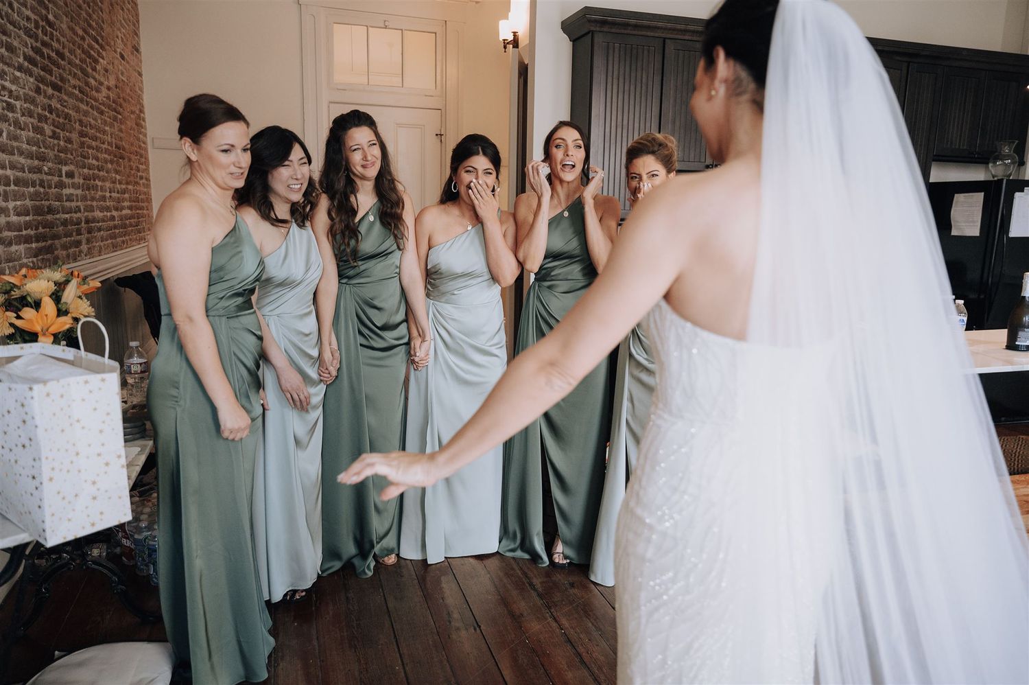 A wedding party in sage green bridesmaid dresses gathers in a rustic brick-walled room.