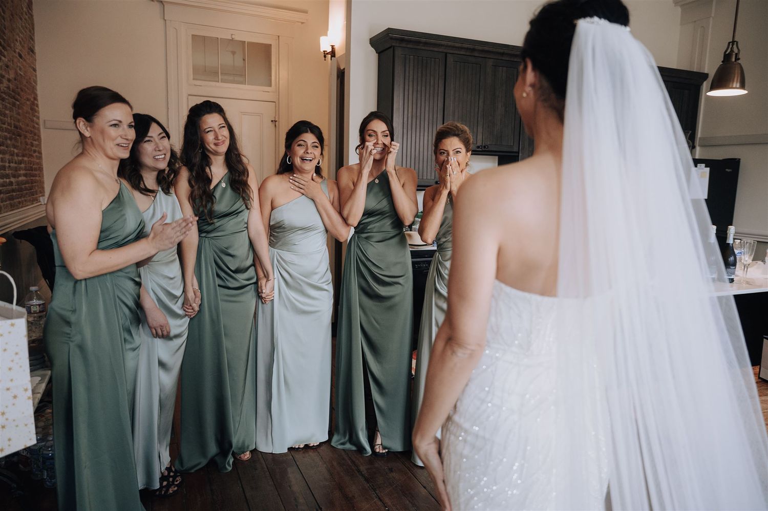 Bridesmaids in matching sage dresses share reactions with bride during pre-wedding preparations.