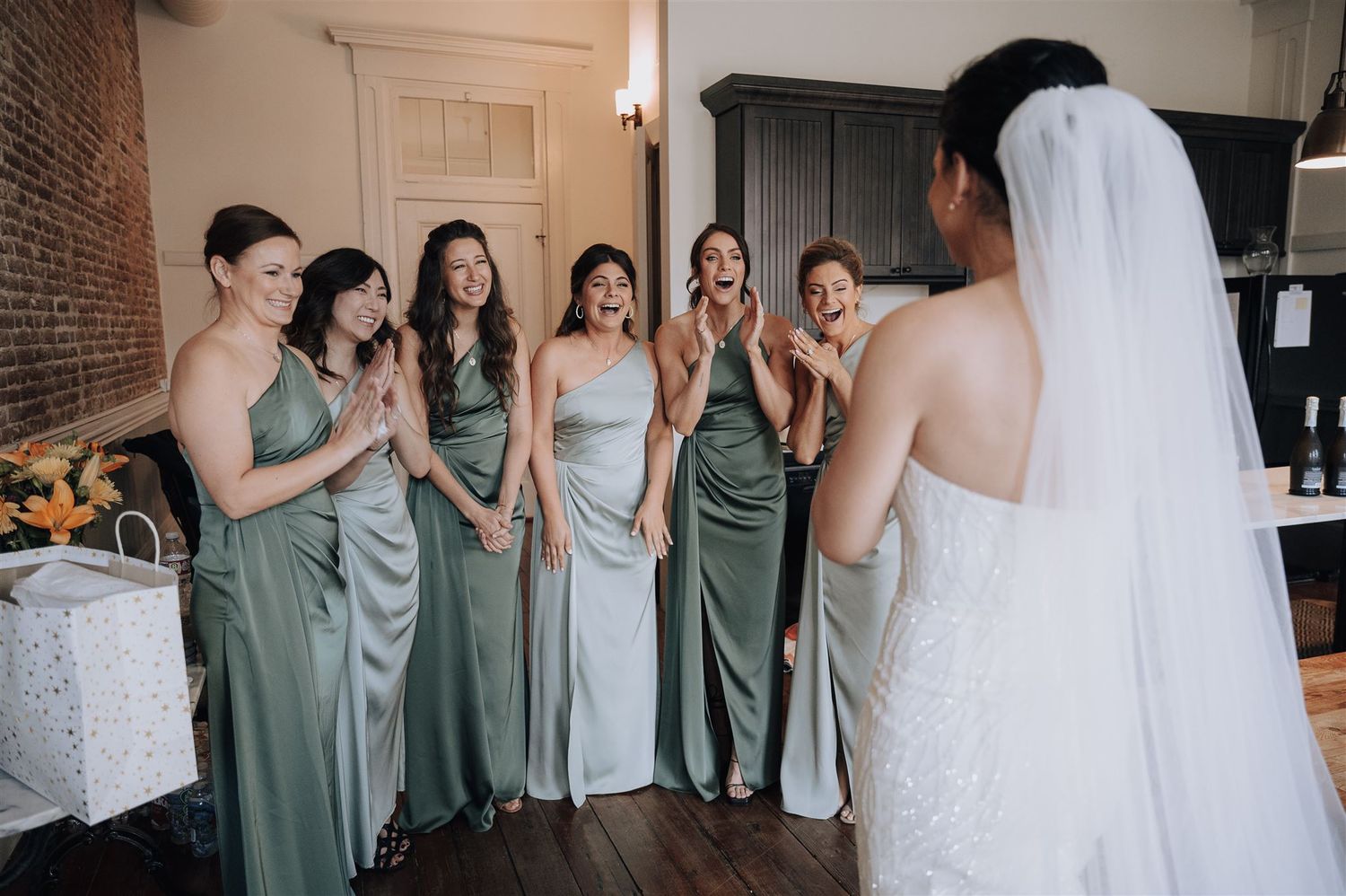 Wedding party in sage green gowns reacts emotionally to bride in white dress and veil.