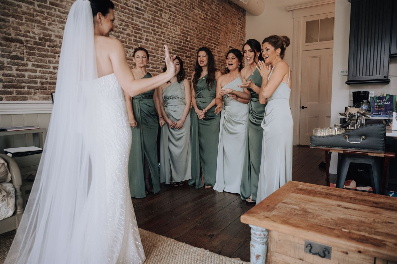 Bride in flowing veil addresses bridesmaids wearing sage green dresses in rustic indoor setting.